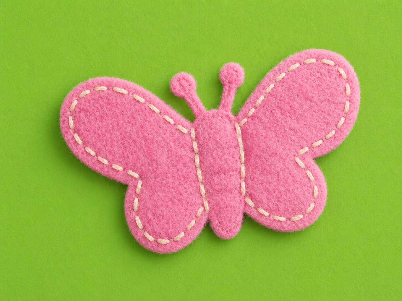 Primer plano de una mariposa de fieltro rosa cosida a mano con hilo blanco, sobre fondo verde.